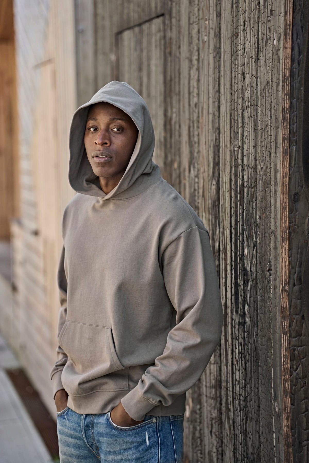 Man wearing the Tee Jays Unlabelled Heavy Loose Fit Hoodie with the hood up, standing by a wooden wall