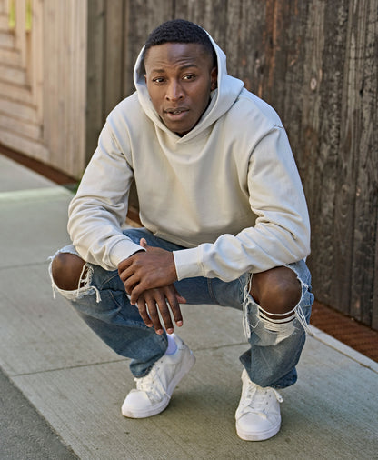 Man wearing the Tee Jays Unlabelled Heavy Loose Fit Hoodie in Cement, crouching on a pavement