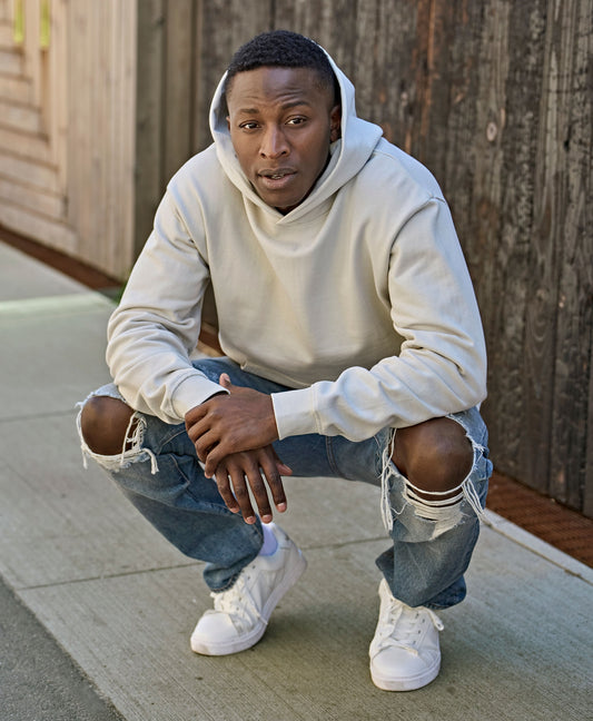 Man wearing the Tee Jays Unlabelled Heavy Loose Fit Hoodie in Cement, crouching on a pavement