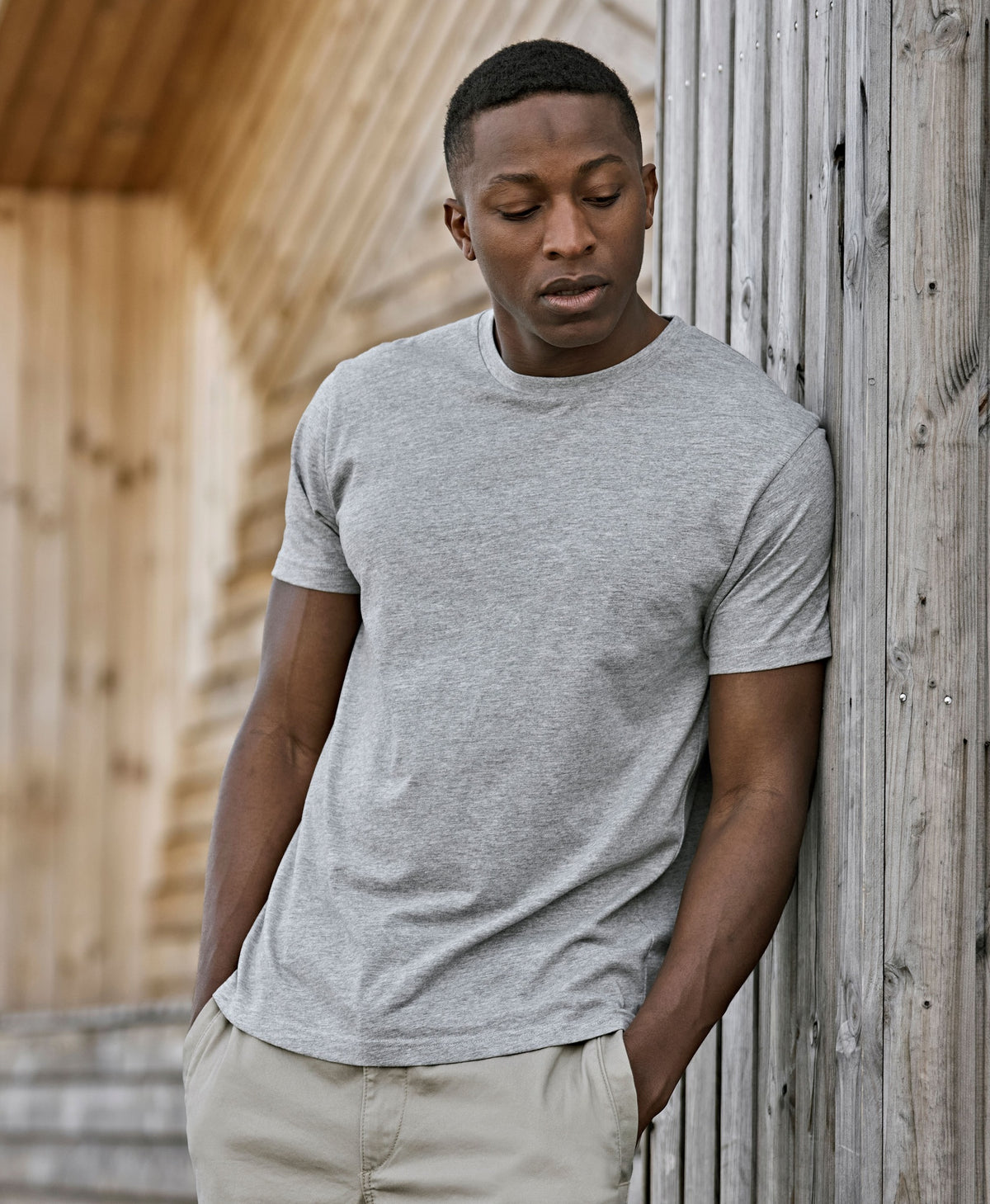 Man wearing the Tee Jays Unlabelled T-Shirt in Heather Grey, leaning on a wooden wall