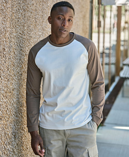 Man wearing the Tee Jays Unlabelled Long Sleeve Baseball T-Shirt in Cement and Clay, standing by a wall