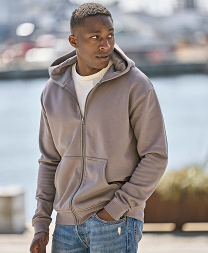 Man wearing the Tee Jays Unlabelled Hooded Full Zip Sweatshirt Hoodie, standing by a harbour