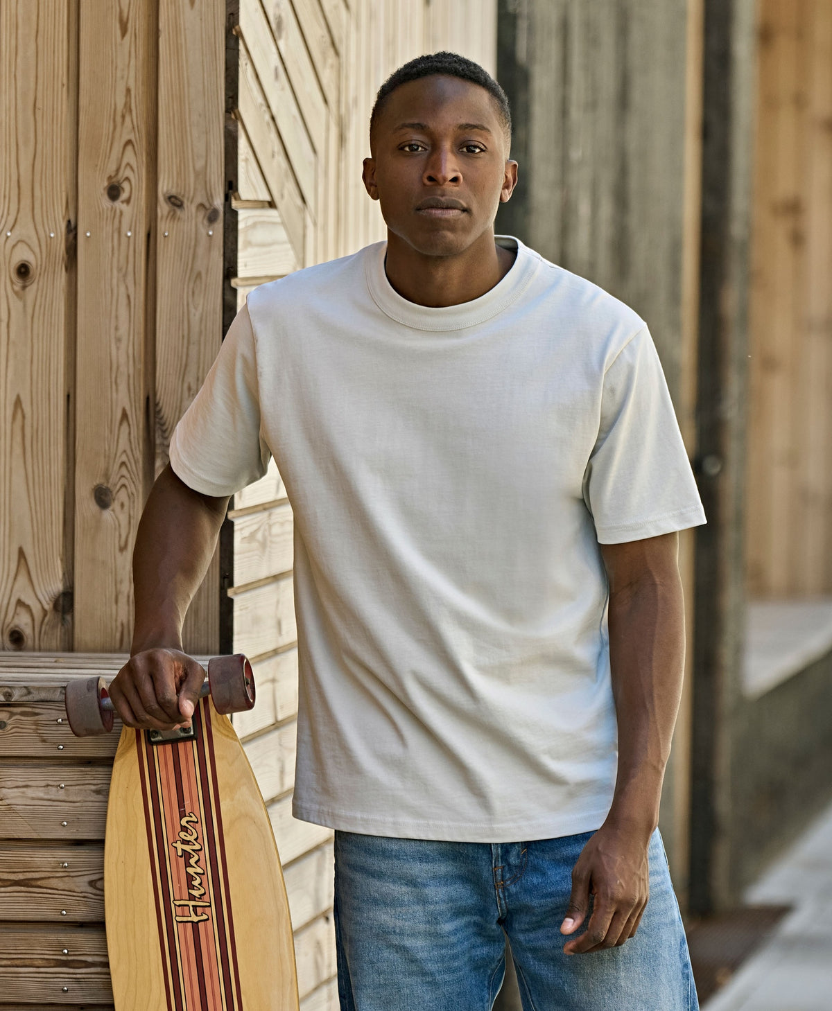 Man wearing the Tee Jays Unlabelled Loose Fit T-Shirt, holding a skateboard by a wooden wall