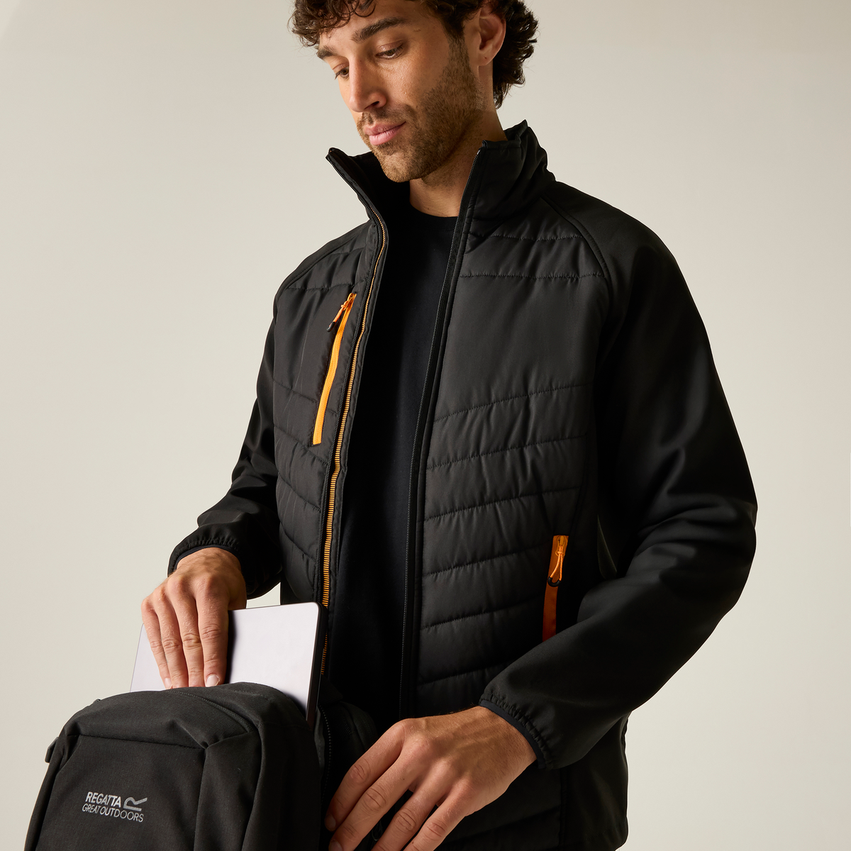 Model wearing the Regatta Pro Men’s Universal Jacket in Black Orange Pop, unzipped, reaching into a bag