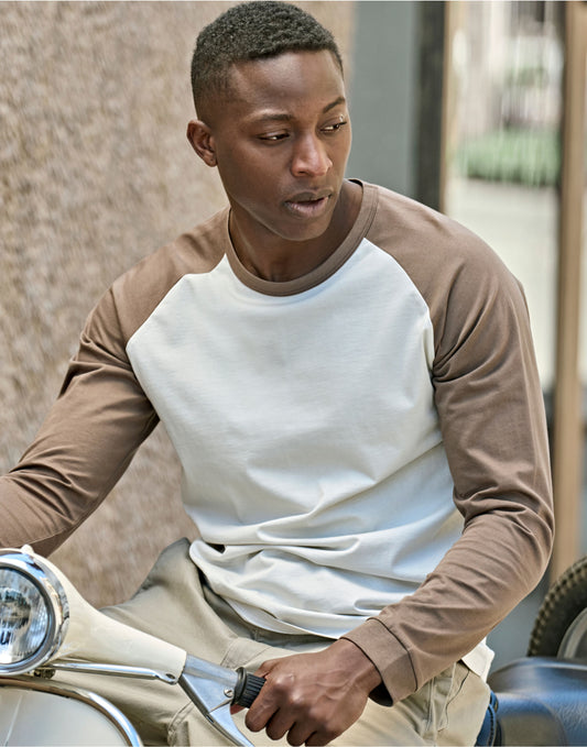 Man wearing the Tee Jays Unlabelled Long Sleeve Baseball T-Shirt in Cement and Clay, leaning on a scooter
