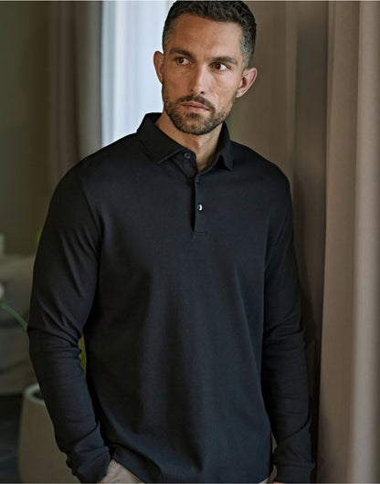 Man wearing the Tee Jays Men’s Pima Cotton Long Sleeve Polo Shirt in Black, standing by a curtain
