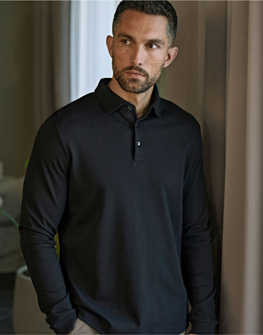 Man wearing the Tee Jays Men’s Pima Cotton Long Sleeve Polo Shirt in Black, standing by a curtain