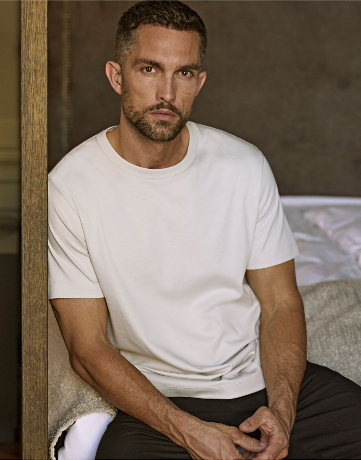 Man wearing the Tee Jays Men’s Pima Cotton T-Shirt, sitting on the edge of a bed