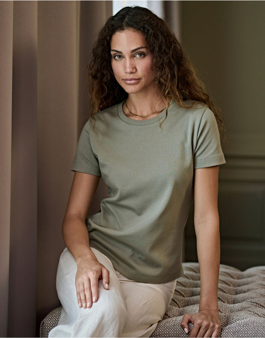 Woman wearing the Tee Jays Ladies Pima Cotton T-Shirt in Sage, sitting on a bed