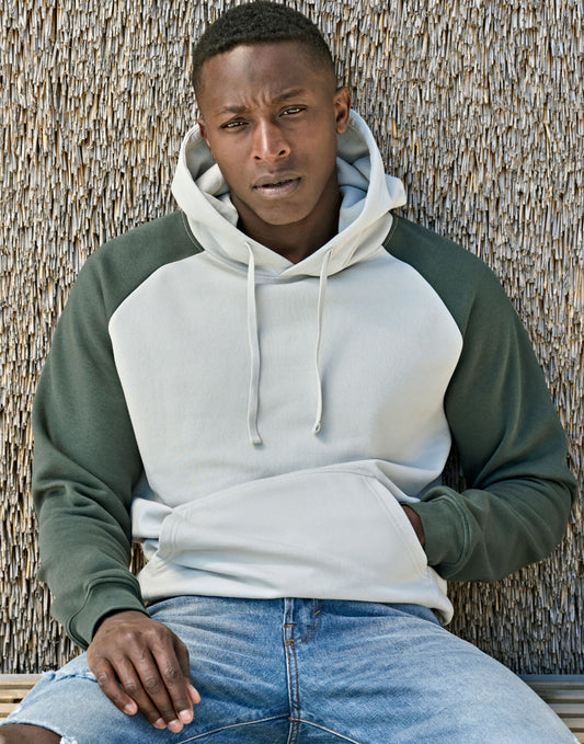 Man wearing the Tee Jays Unlabelled Two-Tone Sweatshirt Hoodie in Cement and Dark Green, sitting against a wall