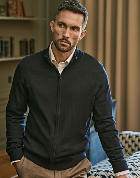 Man wearing the Tee Jays Men’s Full Zip Knit Jumper in Black, sitting in a room with bookshelves