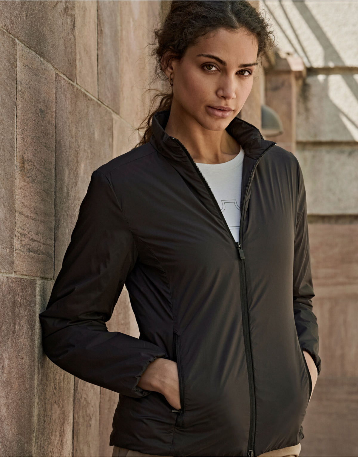 Woman wearing the Tee Jays Ladies PureLite Jacket in Black, standing by a stone wall