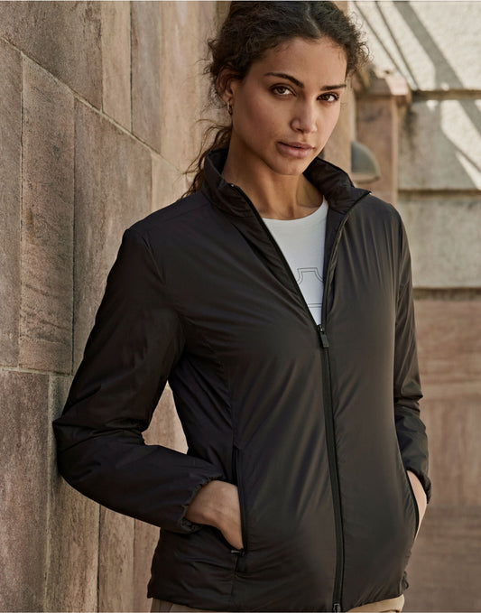 Woman wearing the Tee Jays Ladies PureLite Jacket in Black, standing by a stone wall