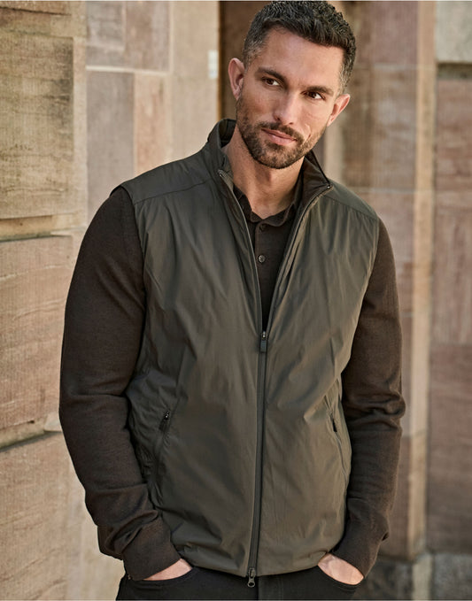 Man wearing the Tee Jays Men’s PureLite Bodywarmer in Shadow, hands in pockets by a stone wall